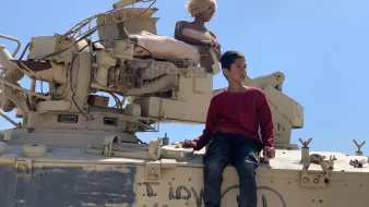 L'image montre un garçon assis sur un véhicule militaire. À côté de lui, il y a une poupée aux cheveux blonds, perchée sur le véhicule. Le fond est clair, avec un ciel bleu. Sur le véhicule, on peut voir des inscriptions, dont "I LOVE YOU" en lettres. L'ensemble de la scène dégage une atmosphere contrastée entre l'innocence de l'enfant et le décor militaire.