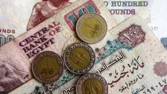 L'image montre des billets et des pièces de monnaie égyptiens. On peut voir un billet de 100 livres égyptiennes, ainsi qu'un billet de 10 livres. En dessous, il y a des pièces de monnaie d'un pound. Les billets sont ornés de motifs et de textes en arabe, indiquant qu'ils proviennent de la Banque Centrale d'Égypte. L'ensemble présente une variété de couleurs et de designs typiques de la monnaie égyptienne.