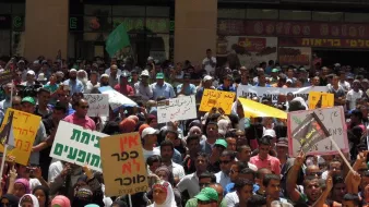 L'image montre une grande foule rassemblée lors d'une manifestation. Les participants tiennent divers panneaux et affiches, certains portant des messages en plusieurs langues, y compris l'hébreu et l'arabe. L'atmosphère semble dynamique, avec des gens exprimant leurs opinions et leurs préoccupations. On peut voir des drapeaux et des bannières, indiquant un fort engagement communautaire et un appel à l'action.