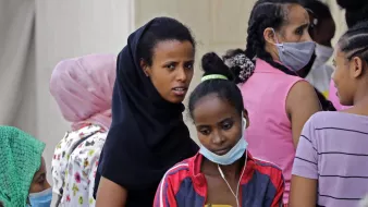 L'image montre un groupe de femmes et de jeunes filles, certaines portant des foulards ou des masques. Les visages expriment divers états d'esprit, allant de la concentration à l'inquiétude. Les vêtements sont colorés, et certaines d'entre elles semblent discuter ou interagir, tandis que d'autres regardent vers l'objectif avec curiosité. L'ambiance générale dénote un moment de rassemblement ou d'attente.