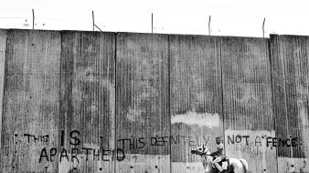 L'image montre un cavalier monté sur un cheval, se tenant près d'un grand mur de béton. Ce mur est marqué par des graffitis en anglais, indiquant des messages provocateurs tels que "THIS IS APARTHEID" et "THIS DEFINITELY NOT A FENCE". L'ambiance de la photo souligne un contraste entre la scène rurale du cavalier et la nature austère du mur, évoquant des thèmes de séparation et de conflit.