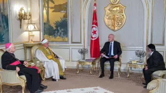 L'image montre une réunion dans un cadre formel. On peut voir plusieurs personnes assises autour d'une table basse. À gauche, un homme en soutane rouge, probablement un évêque, et un homme en robes traditionnelles, au centre, qui semble être un chef religieux. À droite, un homme en costume noir. Au fond, le drapeau tunisien flotte, et on peut apercevoir des éléments de décoration élégants sur les murs et des plantes en arrière-plan. La scène évoque un moment de dialogue interreligieux ou de rencontre officielle.