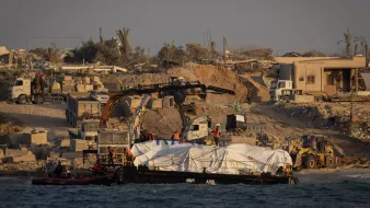 L'image montre une scène de construction ou de travaux sur une côte. On peut voir un bateau sur l'eau avec une grande bâche blanche, probablement transportant des matériaux ou des fournitures. Sur la rive, il y a des machines de construction, comme des pelleteuses et des camions, ainsi que des travailleurs en action. En arrière-plan, des bâtiments ruinés et des palmiers suggèrent un environnement en mutation, peut-être en raison de travaux de développement ou de reconstruction. L'atmosphère générale semble celle d'un chantier en cours.