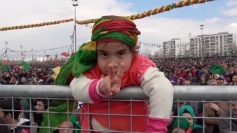 L'image montre une jeune fille participant à un grand rassemblement. Elle est vêtue de vêtements colorés, portant un foulard aux teintes vives. La fillette fait un signe de paix avec ses doigts, pendant qu'elle se penche sur une barrière. En arrière-plan, une grande foule est présente, témoignant d'un événement festif ou d'une manifestation populaire, avec des décorations colorées suspendues au-dessus. L'ambiance semble joyeuse et engagée.