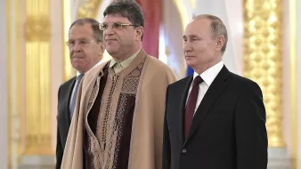 L'image montre trois hommes en pleine cérémonie. Au centre, un homme porte une tenue traditionnelle ornée de motifs. À sa gauche, un homme en costume formel et à sa droite, un homme aussi en costume. Le décor semble somptueux avec des dorures et des drapeaux en arrière-plan, suggérant un lieu officiel. L'atmosphère semble sérieuse et formelle.
