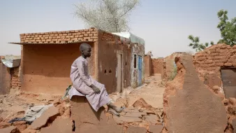 L'image montre un jeune garçon assis sur de décombres dans une région désertique. Il porte une tunique traditionnelle et semble pensif. En arrière-plan, il y a des bâtiments en terre cuite, dont certains sont partiellement détruits. Le paysage est aride, avec quelques arbres éparpillés, et le ciel est clair. L'atmosphère dégage une impression de solitude et de résilience face aux conditions difficiles.