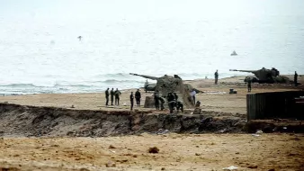 L'image montre une scène côtière où des militaires sont visibles sur une plage. On aperçoit des canons posés sur le sable, ainsi qu'un groupe de soldats qui semblent s'affairer autour. Le paysage est brumeux, avec une mer calme au fond et un ciel nuageux. L'atmosphère paraît sérieuse et militaire, avec une concentration sur les préparatifs ou les opérations en cours.