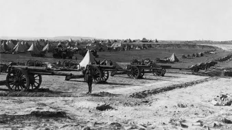 L'image montre un camp militaire avec plusieurs canons en premier plan. À l'arrière-plan, on distingue des tentes disposées de manière ordonnée sur un terrain dégagé. Le paysage semble vaste, avec des collines ou des montagnes au loin. L'atmosphère est historique, évoquant une période de guerre ou de mobilisation des troupes. Un soldat ou un homme se tient près des canons, donnant une échelle à la scène.