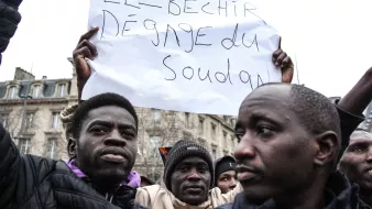 L'image montre une manifestation où des personnes tiennent des pancartes. L'une d'elles est clairement visible et affiche un message en français disant "El-Béchir dégage du Soudan". Les participants semblent passionnés et engagés dans une cause. On peut discerner des expressions sérieuses et déterminées sur leurs visages. En arrière-plan, des bâtiments peuvent être aperçus, suggérant un cadre urbain.