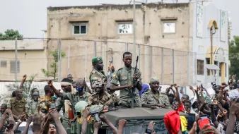 L'image montre un groupe de soldats, armés, sur un véhicule militaire, entourés par une foule enthousiaste. Les soldats semblent célébrer ou soutenir une cause, tandis que les membres de la foule lèvent les bras et expriment leur joie ou leur enthousiasme. En arrière-plan, on peut apercevoir des bâtiments et une atmosphère de mobilisation populaire.