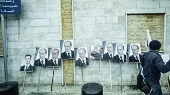 L'image montre un mur en béton avec plusieurs portraits accrochés, principalement des visages d'un homme en costume. Ces portraits sont placés sur des supports en bois. Une personne, dont on ne voit que le dos, marche à proximité. Le décor semble urbain et pourrait impliquer un contexte politique. Les expressions faciales sur les portraits sont sérieuses, reflétant une ambiance de propagande ou de soutien.