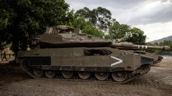 L'image montre un char d'assaut stationné sur un terrain. Le véhicule est de couleur verte foncée et possède des marques de camouflages. Il est équipé d'une tourelle avec un canon pointé vers l'avant. En arrière-plan, on peut voir des arbres et un ciel nuageux, ce qui donne une ambiance militaire à la scène. Le char semble robuste et bien conservé, prêt pour une utilisation éventuelle.