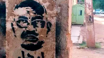 L'image montre un graffiti sur un mur, représentant un visage stylisé d'un homme avec des lunettes. Le dessin est en noir, sur un fond de béton clair. En arabe, le mot "ارحل" est inscrit, ce qui signifie "pars" ou "sors". L'environnement semble urbain, avec des bâtiments et une route visible en arrière-plan.
