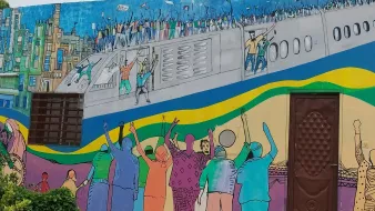L'image montre une fresque murale colorée qui illustre une scène dynamique. On y voit des personnes célébrant et manifestant, les bras levés, sur un fond représentant un avion et une ville. Les couleurs vives ajoutent une énergie palpable à la scène, reflétant un sentiment de joie ou de solidarité. Des silhouettes de différents personnages sont visibles, et l'ensemble de l'œuvre évoque un message de rassemblement et d'espoir.