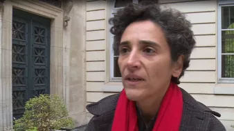 L'image montre une femme avec des cheveux bouclés, portant un manteau sombre et une écharpe rouge. Elle semble s'exprimer avec sérieux, peut-être dans le cadre d'une interview. En arrière-plan, on aperçoit un bâtiment avec des fenêtres et une porte d'entrée, ce qui donne une impression urbaine et architecturale.