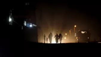 L'image montre un groupe de personnes marchant sur une route sombre, illuminée par des lumières vives au loin. Les silhouettes des individus sont visibles, créant une ambiance mystérieuse. En arrière-plan, on aperçoit des bâtiments et d'autres sources de lumière, suggérant un environnement urbain. L'atmosphère est à la fois sombre et intrigante, mettant en avant le contraste entre l'ombre et la lumière.