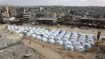 L'image montre une vaste zone de tentes blanches installées sur un terrain aride, entourée de bâtiments en ruines. Les tentes semblent former un camp, probablement pour des personnes déplacées ou des réfugiés. En arrière-plan, on peut voir des structures détruites, ce qui indique un environnement de conflit ou de catastrophe. Le ciel est nuageux, et l'atmosphère générale évoque la désolation et la nécessité d'abri.