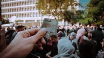 L'image montre une main tenant un billet de banque marocain au premier plan, tandis qu'à l'arrière-plan, une foule est rassemblée. On aperçoit des personnes aux tenues variées, certaines portant des foulards. Le cadre semble être un événement public, probablement une manifestation ou une célébration, avec des bâtiments modernes en arrière-plan et des arbres qui ajoutent une touche de verdure à la scène. L'atmosphère semble dynamique et chargée d'énergie collective.