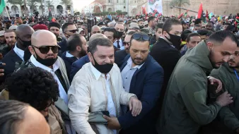 L'image montre une foule dense, avec un groupe de personnes rassemblées lors d'un événement. Au centre, un homme en tenue traditionnelle est entouré de plusieurs personnes portant des masques. L'ambiance semble être celle d'un rassemblement ou d'une célébration. Les participants affichent des expressions d'enthousiasme, certains tenant des bannières ou des drapeaux. En arrière-plan, des bâtiments et de la verdure sont visibles, ce qui indique une atmosphère festive.