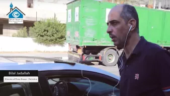 L'image présente un homme marchant à côté d'une voiture, portant des écouteurs. Il a un air concentré et semble être dans un environnement urbain avec un camion vert en arrière-plan. Un texte en bas de l'image mentionne "Bilal Jadallah, Directeur de Press House - Palestine".