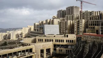 L'image montre un paysage urbain en construction, avec des bâtiments modernes de forme géométrique et des structures encore inachevées. On peut voir des façades en béton et des fenêtres non terminées sur plusieurs des bâtiments. En arrière-plan, une grue rouge indique que des travaux sont en cours. Le ciel est nuageux, créant une atmosphère un peu sombre. Au premier plan, des éléments de construction sont visibles, suggestifs d'un site en développement.