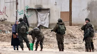 L'image montre une femme vêtue d'une robe sombre, se tenant à côté d'une rue. Devant elle, plusieurs soldats en uniforme sont regroupés, certains sont en train de fouiller un sac, tandis que d'autres observent la femme. En arrière-plan, on peut voir un mur avec des graffitis et des débris sur le sol.