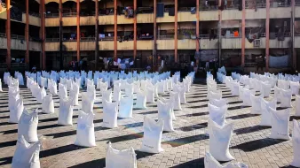 L'image représente un espace ouvert où de nombreux sacs blancs sont disposés en lignes. Ces sacs semblent être remplis de quelque chose, et le sol est constitué de pavés. En arrière-plan, on peut voir un bâtiment à plusieurs étages avec des balcons. Des vêtements sont suspendus à l'extérieur des balcons, suggérant une utilisation résidentielle de cet espace. La lumière du soleil éclaire la scène, créant un contraste entre les sacs blancs et l'environnement.