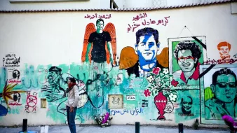 L'image montre un mur recouvert de fresques murales colorées. On y voit plusieurs portraits et illustrations. L'une des figures centrales semble représenter une personne avec des ailes, symbole souvent associé à un message d'espoir ou de mémoire. Des textes en arabe et en français peuvent être aperçus, évoquant des thèmes de la liberté et de la résistance. En bas, une personne se tient debout, probablement en train de peindre ou d'admirer l'œuvre. L'atmosphère générale dégage une impression vibrante et engagée, reflétant une culture urbaine riche.
