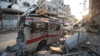 L'image montre une scène de destruction urbaine, probablement à la suite d'un conflit. On y voit une ambulance endommagée, avec sa structure déformée et son revêtement extérieur marqué par des débris. Les bâtiments environnants sont en ruine, avec des fenêtres brisées et des murs partiellement effondrés. Le sol est jonché de débris, et l'éclairage indique que c'est soit tôt le matin, soit en fin d'après-midi. L'atmosphère générale est sombre et témoigne de l'impact des violences sur la ville.