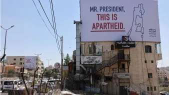 L'image montre un panneau publicitaire accroché sur un bâtiment, avec un message qui dit "MR. PRESIDENT, THIS IS APARTHEID." On peut voir des infrastructures urbaines environnantes, telles que des routes, des bâtiments et des fils de téléphone. Le contexte suggère une critique sociale ou politique liée à la situation en Israël-Palestine. Le paysage est ensoleillé et présente une ambiance de ville.