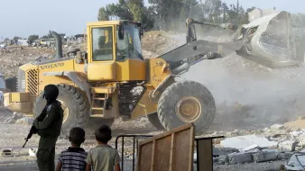 L'image montre un camion-tracteur de type Volvo en action, probablement en train de déblayer ou de détruire des structures. En premier plan, deux jeunes enfants observent la scène. À l'arrière-plan, on peut voir des débris et des bâtiments partiellement détruits, ainsi qu'un soldat en tenue militaire. L'atmosphère semble tendue, avec de la poussière soulevée par le véhicule.