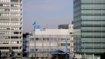 L'image montre le bâtiment de l'OPEP (Organisation des pays exportateurs de pétrole), qui est un bâtiment moderne à plusieurs étages. Des drapeaux sont visibles sur le toit, y compris celui de l'OPEP. Le bâtiment est entouré par d'autres structures urbaines, reflétant un environnement de ville. Le ciel est légèrement nuageux, suggérant une journée calme. Des éléments architecturaux en verre et en béton contrastent avec le design du bâtiment de l'OPEP.