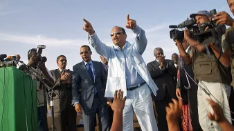 L'image montre un homme debout sur une scène, habillé en costume clair. Il semble être en train de faire un discours et de pointer vers le public. Autour de lui, plusieurs personnes l'écoutent attentivement, dont certaines sont en train de prendre des photos ou de filmer avec des caméras. Il y a une ambiance d'événement public ou politique, avec un discours énergique et engageant.