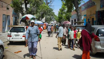 L'image montre une rue animée avec des groupes de personnes marchant. On peut voir un homme âgé utilisant une canne, habillé en bleu, qui semble avancer lentement. Plusieurs personnes sont dispersées le long de la rue, certaines se trouvent près de voitures garées. Des ombrelles colorées sont visibles, probablement pour se protéger du soleil. Les bâtiments en arrière-plan semblent être de petits commerces ou habitations. L'ambiance générale dénote une vie quotidienne dynamique dans un environnement urbain.
