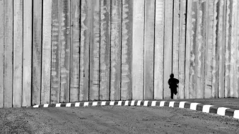 L'image montre un enfant courant le long d'un mur de béton. Le mur est haut et semble imposant, créant une ambiance de séparation ou d'isolement. Le contraste en noir et blanc accentue les textures du mur et la silhouette de l'enfant, qui semble minuscule par rapport à la structure massive. Le sol est désertique avec une route qui tourne légèrement, ajoutant à l'impression de solitude et de mouvement.