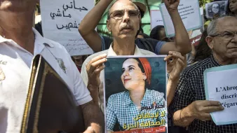 L'image montre un groupe de personnes rassemblées pour une manifestation. Au premier plan, un homme tient une affiche qui représente une femme avec un sourire, probablement une figure symbolique pour le mouvement. Les participants, majoritairement des adultes, tiennent également des pancartes avec des slogans en arabe et en français, indiquant leur lutte ou leur soutien à une cause, sans doute liée à des droits ou à une injustice. L'atmosphère semble déterminée et engagée.