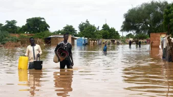L'image montre une scène de inondation où deux hommes marchent dans une zone inondée. L'eau monte jusqu'à leurs jambes et environ une partie des maisons environnantes est submergée. Les hommes portent des objets, l'un avec un seau et l'autre avec des paniers ou des matelas. En arrière-plan, on peut voir des arbres et des bâtiments partiellement immergés, représentant une situation difficile due aux inondations. Le ciel est nuageux, ce qui suggère des conditions météorologiques instables.