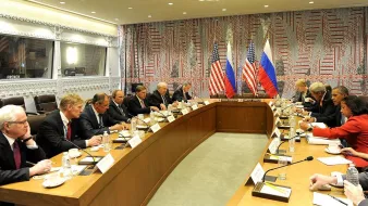 L'image montre une réunion formelle se déroulant dans une salle de conférence. À une longue table, des représentants des États-Unis et de la Russie sont présents, avec des drapeaux des deux pays visibles en arrière-plan. Les participants, qui semblent être des diplomates ou des responsables gouvernementaux, sont engagés dans une discussion sérieuse. La décoration de la salle présente des motifs géométriques sur les murs, et l'ambiance générale est celle d'une négociation ou d'un dialogue diplomatique.