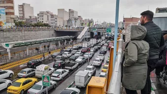 L'image montre une scène de circulation dense dans une ville. On peut voir de nombreuses voitures de différentes marques immobilisées dans un embouteillage. À l'arrière-plan, on distingue des bâtiments urbains et une route à plusieurs niveaux. Il y a des panneaux de signalisation visibles, et deux personnes se tiennent sur un balcon ou une balustrade, observant la circulation en contrebas. Le ciel est nuageux, ce qui suggère un temps gris et éventuellement pluvieux.