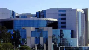 L'image montre un bâtiment moderne au design architectural contemporain. La façade est principalement en verre, ce qui reflète la lumière et les bâtiments environnants. Des formes géométriques et des courbes sont bien mises en valeur, et on peut distinguer des éléments en métal. Le paysage autour est agrémenté d'arbres et de plantes, suggérant que le bâtiment est situé dans une zone urbaine. L'ensemble dégage une impression de sophistication et de modernité.