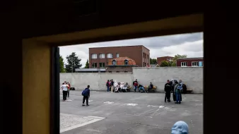 L'image montre une scène d'un espace extérieur, probablement une cour de récréation ou un terrain de sport. On peut voir plusieurs groupes de jeunes, répartis sur le sol, assis ou discutant ensemble. À l'arrière-plan, il y a des bâtiments avec des fenêtres et un toit de briques. Le ciel est nuageux, ce qui donne une ambiance un peu sombre à la scène. Les jeunes semblent engagés dans des conversations ou des activités sociales.