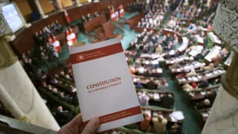L'image montre une scène à l'intérieur d'une assemblée, probablement un parlement, avec un grand nombre de personnes assises dans une salle. Au premier plan, on voit une personne tenant un document portant le titre "CONSTITUTION DE LA RÉPUBLIQUE TUNISIENNE". L'assemblée est ornée de drapeaux, et l'ambiance semble solennelle, suggérant un moment important lié à la constitution ou à la législation.