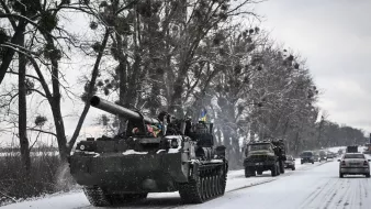 L'image montre un paysage hivernal avec de la neige recouvrant le sol. Un char militaire, probablement un tank, avance sur une route étroite bordée d'arbres. Des véhicules militaires sont également visibles en arrière-plan, créant une impression de mouvement dans un environnement froid et désert. L'atmosphère est marquée par des nuances de gris et de blanc, typiques d'un paysage d'hiver.