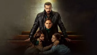 L'image présente une scène dramatique avec quatre personnages. Au centre, une femme est assise, entourée de deux jeunes filles qui semblent chercher protection. La femme a une expression préoccupée, tandis que l'homme derrière elle, vêtu d'une veste en cuir, dégage une présence protectrice et intense. L'éclairage crée un contraste marqué, accentuant l'atmosphère tendue et émotive de la scène. Les éléments de l'environnement, comme les bancs, suggèrent un cadre intérieur, peut-être une église ou un endroit similaire, renforçant le caractère sérieux de la situation.