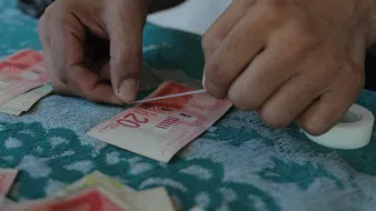 L'image montre des mains qui manipulent des billets de banque. Une personne semble être en train de recoller ou d'ajuster un billet de 20 unités. On peut voir un surface en tissu avec des motifs, ainsi que d'autres billets éparpillés autour. La situation donne une impression de travail minutieux avec une attention particulière aux détails.