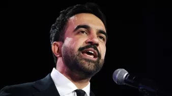 Homme en costume noir, parlant au micro avec une expression engagée.