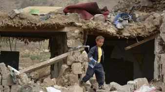 L'image montre un enfant marchant à travers des décombres d'un bâtiment détruit. Les murs sont effondrés et de nombreux débris, tels que des pierres et des objets éparpillés, sont visibles autour de lui. L'environnement semble être celui d'une zone touchée par un désastre, avec un paysage en arrière-plan qui pourrait suggérer des conditions difficiles. L'enfant porte des vêtements sombres, et son expression évoque une certaine gravité face à la situation environnante.