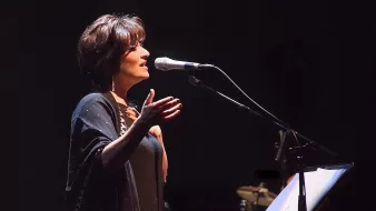 L'image montre une femme sur scène, en train de chanter ou de parler dans un microphone. Elle a les cheveux courts et bruns, et porte une tenue sombre, accompagnée d'un châle. Son expression semble engagée et passionnée, suggérant qu'elle interprète une performance émotionnelle. En arrière-plan, on aperçoit des instruments de musique, ce qui indique qu'elle se produit dans un cadre musical. L'éclairage est tamisé, créant une ambiance intime et captivante.