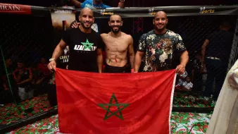 L'image montre trois hommes posant ensemble à l'intérieur d'un environnement de combat, probablement après une victoire. L'un d'eux est torse nu et affiche un large sourire, tandis que les autres sont habillés avec des t-shirts. Ils tiennent une grande drapeau marocain, mettant en avant leur fierté nationale. Le sol est couvert de confettis, ajoutant une atmosphère festive à la scène. En arrière-plan, on peut voir des spectateurs soulevant les bras en signe de célébration.