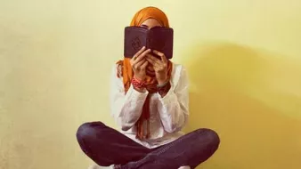 L'image montre une personne assise en tailleur, tenant un livre près de son visage, cachant ainsi son identité. Elle porte un hijab orange et une tenue simple, avec des jeans. L'arrière-plan est d'une couleur claire, créant une ambiance douce et calme. Les bras sont repliés et des bracelets colorés ornent ses poignets. Cette image évoque un moment de réflexion ou de lecture.